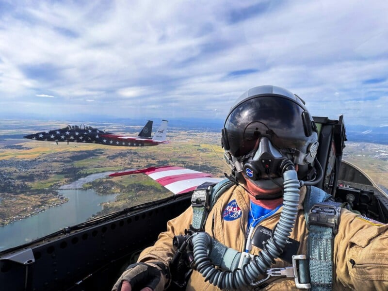 A pilot in a flight suit and helmet takes a selfie inside a jet cockpit while another jet, decorated with a stars and stripes design, flies nearby over a scenic landscape with water and fields.