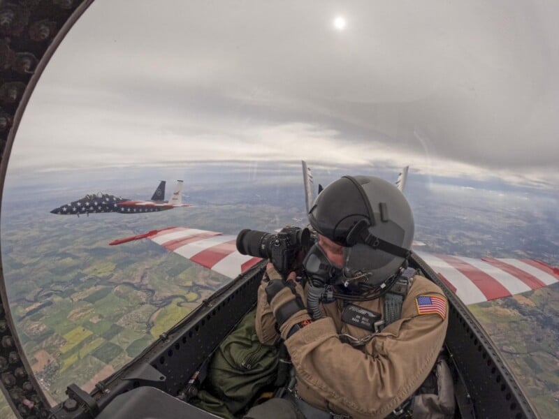 A pilot in a cockpit takes photos with a camera, wearing a helmet and flight suit. Another jet, decorated with a U.S. flag motif, flies nearby above a landscape of fields and clouds.