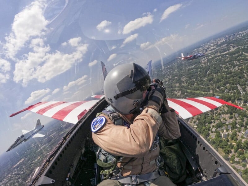 A pilot in a jet with red-and-white striped wings takes a photo mid-flight; another jet and scenery with houses and trees are visible below under a partly cloudy sky.