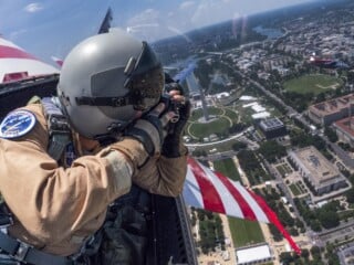 A person in a flight suit and helmet takes photos from an aircraft above Washington, D.C., with the Washington Monument and cityscape visible below. An American flag pattern is seen on the aircraft’s wing.