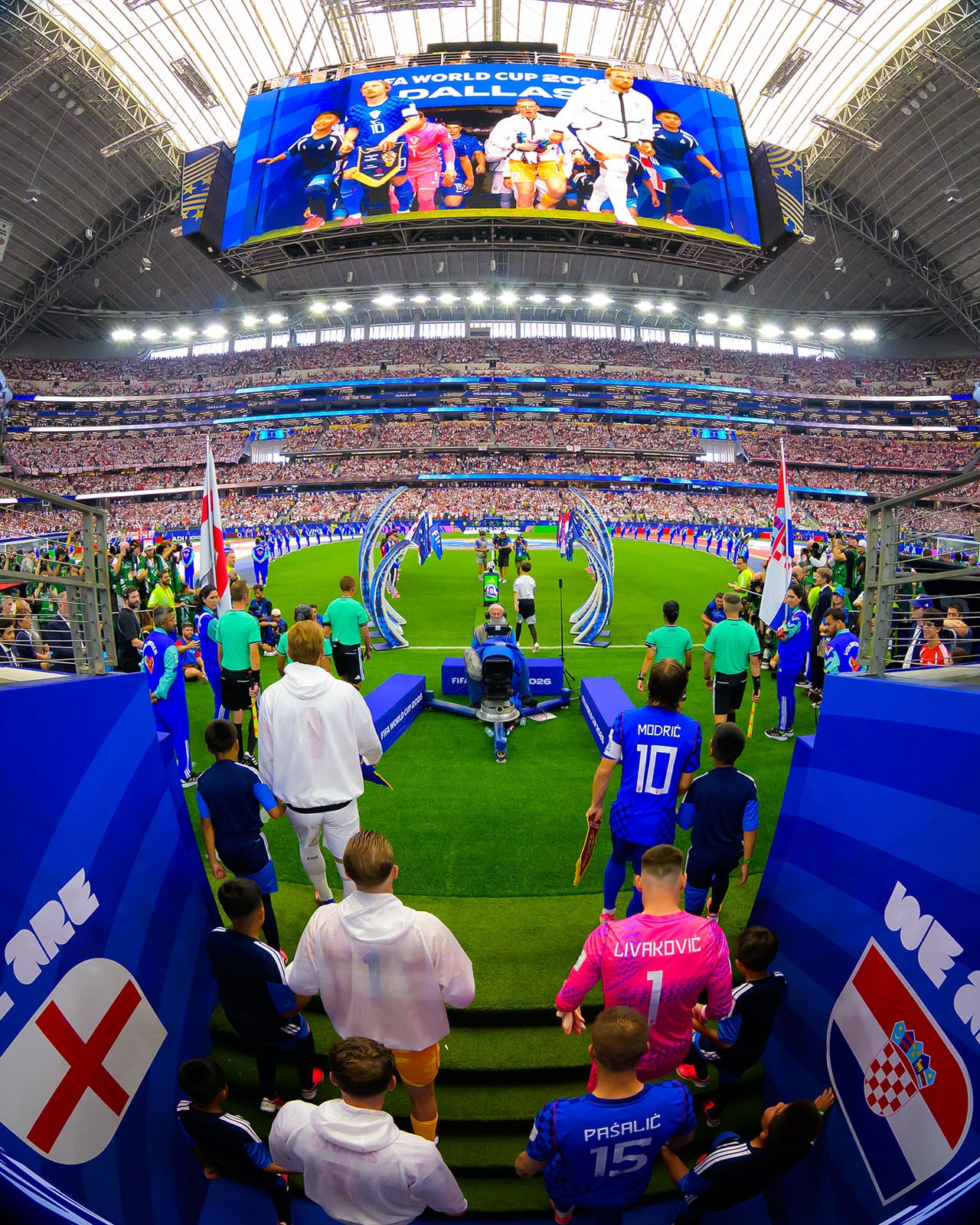 Soccer players from England and Croatia walk onto the field in a packed stadium, with fans watching and flags displayed. Bright lights illuminate the indoor arena.