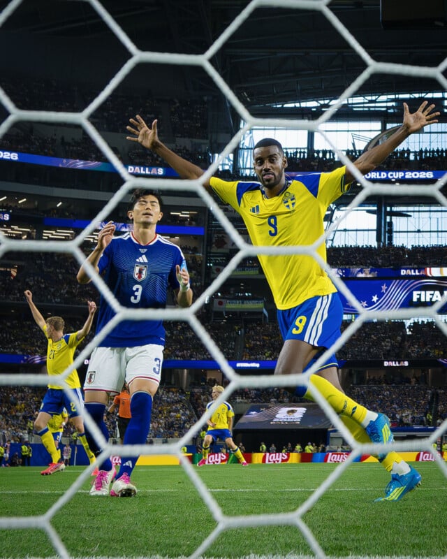A soccer player in a yellow Sweden jersey celebrates a goal with arms raised, while a Japanese defender reacts nearby. The scene is viewed through the net in a crowded stadium.