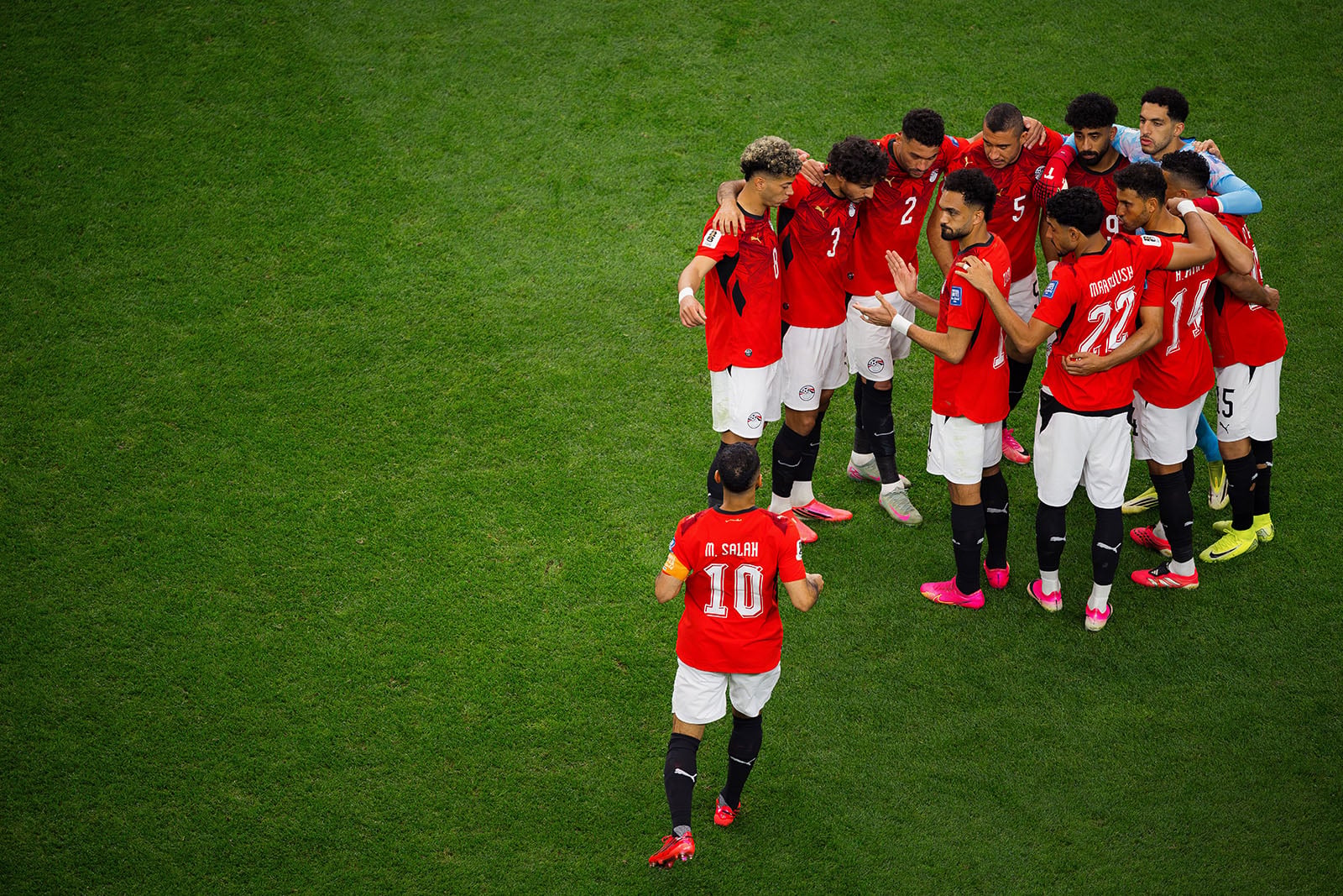 A soccer team in red jerseys huddles on the field, while one player wearing number 10 approaches to join them. Green grass covers the background.