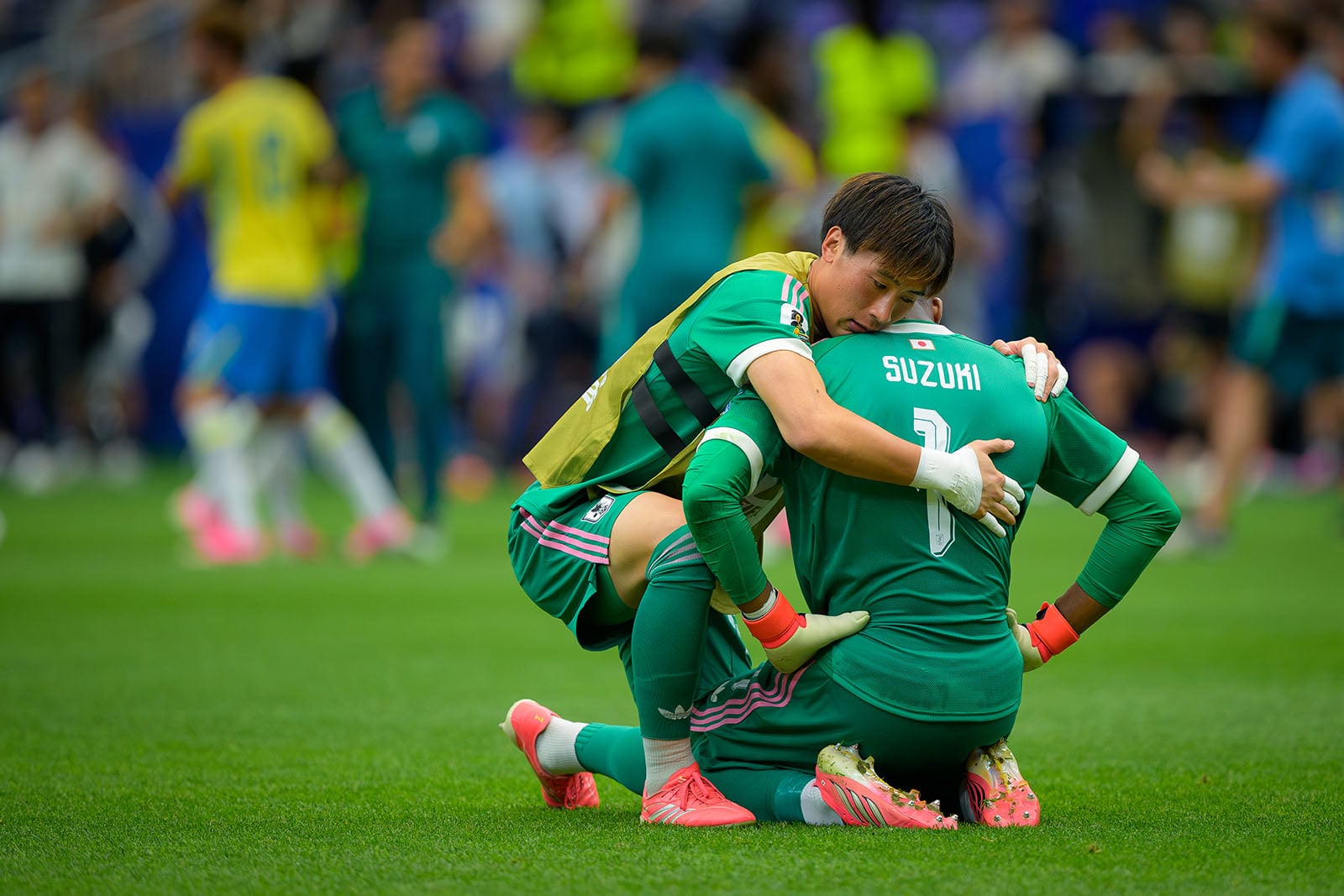 Two soccer players in green uniforms embrace on the field, one comforting the other who is kneeling with "SUZUKI 1" on their back. The scene suggests a moment of consolation after a match.