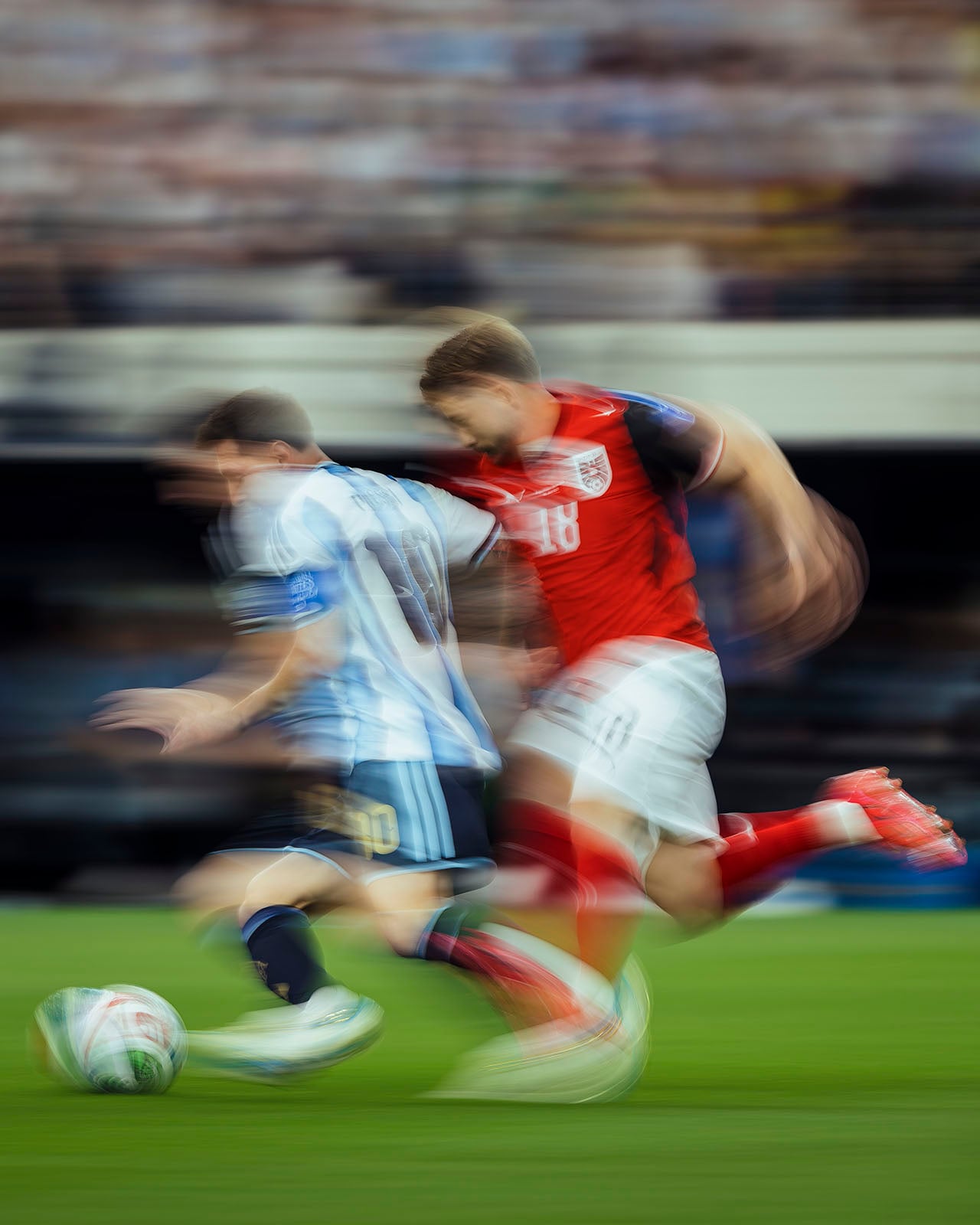 Two soccer players in motion blur, one in a blue and white striped jersey and the other in a red jersey, compete for the ball on a green field during a match.