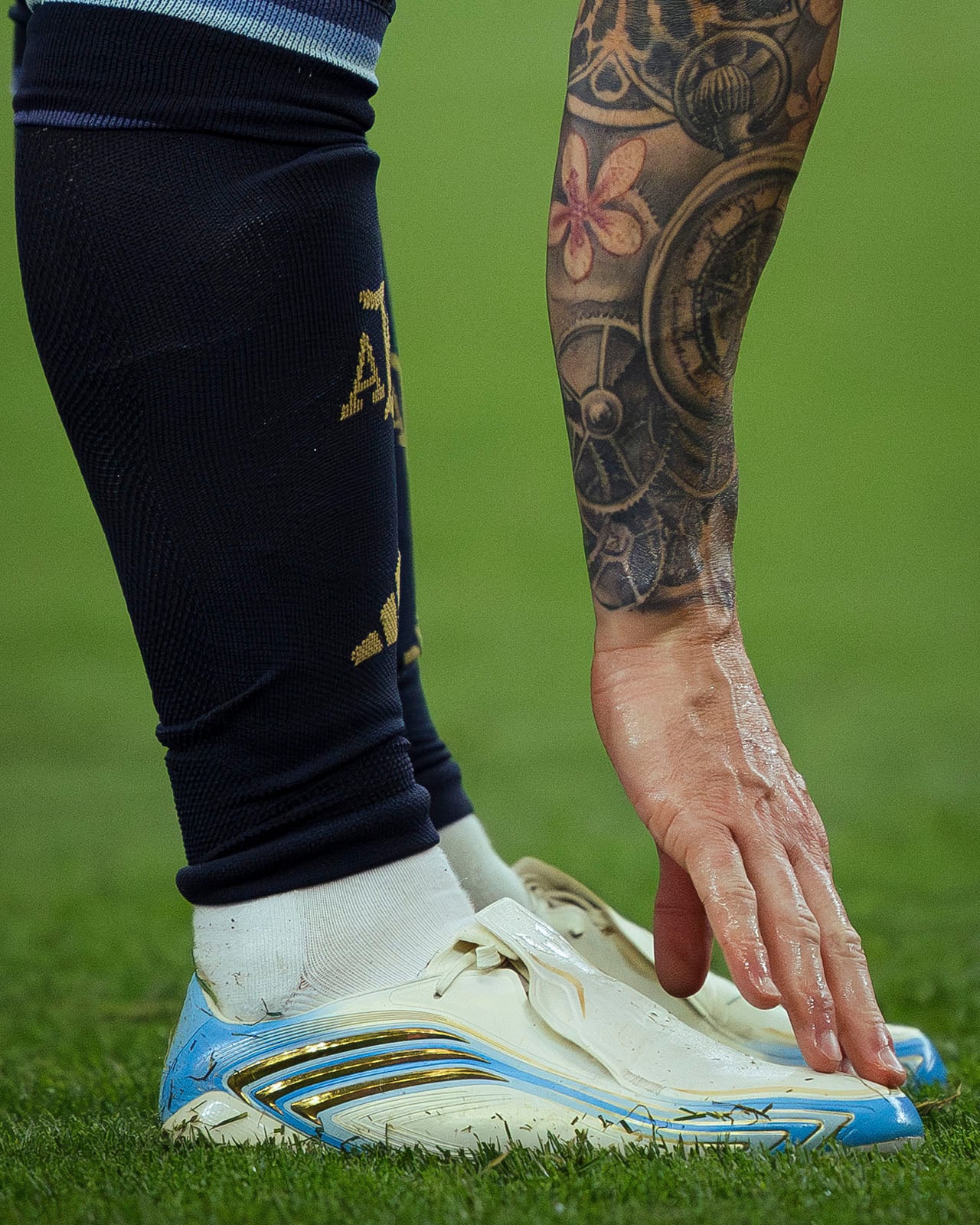 A tattooed arm reaches down toward white and blue soccer cleats on grass. The person wears dark socks featuring yellow lettering and graphics.
