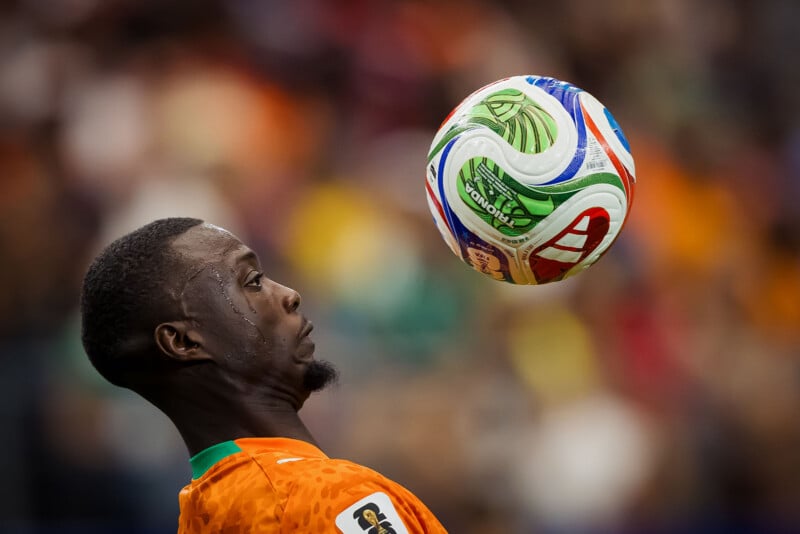 A soccer player in an orange jersey focuses on a colorful ball in mid-air, preparing to control it with his chest. The background is blurred, highlighting the player's concentration.
