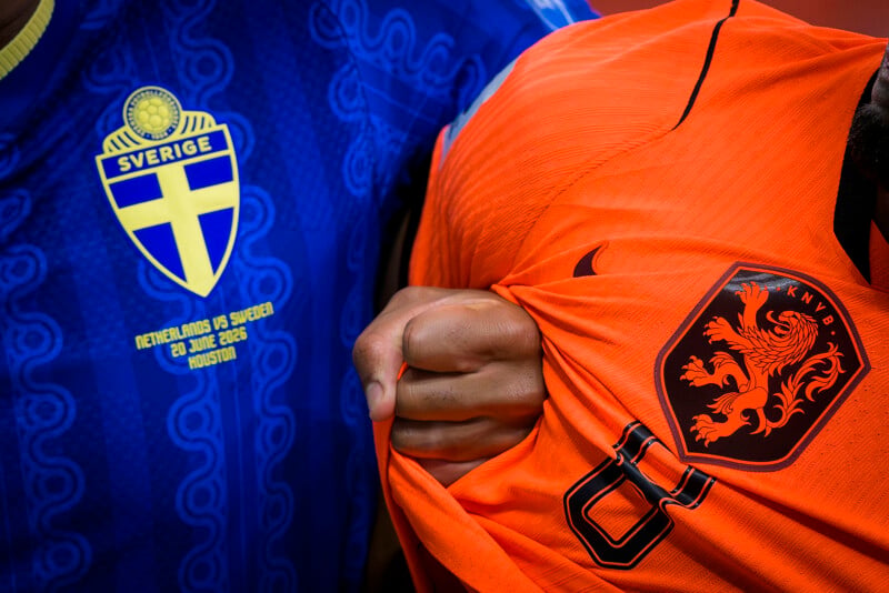 A close-up of a Swedish soccer jersey next to a Dutch soccer jersey, with a hand gripping the Dutch jersey. Both have team logos and match information visible.