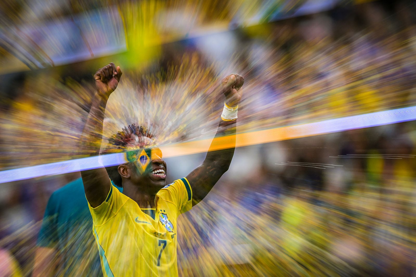 A soccer player in a yellow Brazil jersey raises both arms in celebration, with a motion blur effect radiating outward, creating a dynamic, energetic scene.