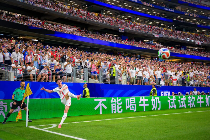 A soccer player kicks a corner ball during a match in a packed stadium, with fans cheering in the stands and referees watching near the field.