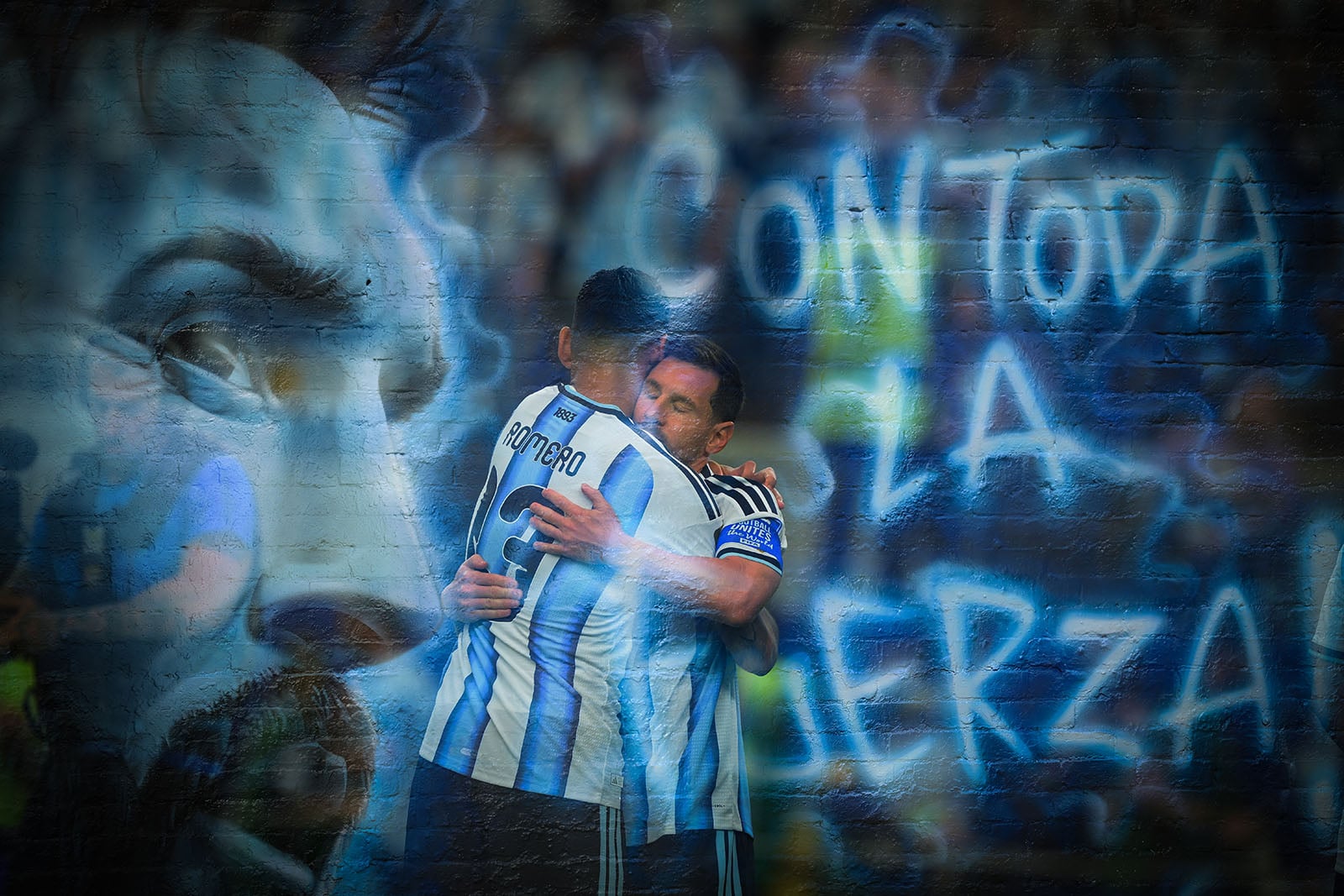 Two soccer players in Argentina jerseys embrace in front of a mural featuring a large face and blue graffiti text reading “Con toda la fuerza” on a brick wall.