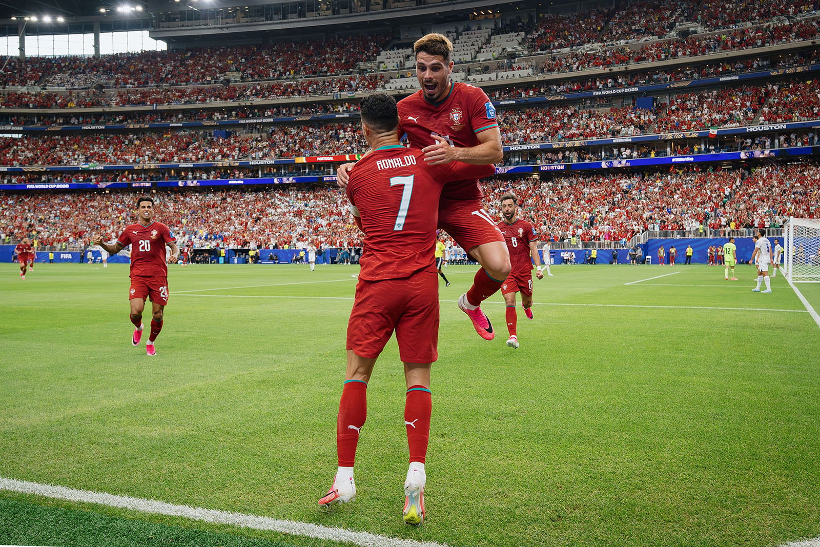 Two soccer players in red uniforms celebrate a goal; one jumps into the other's arms. A packed stadium with cheering fans fills the background.