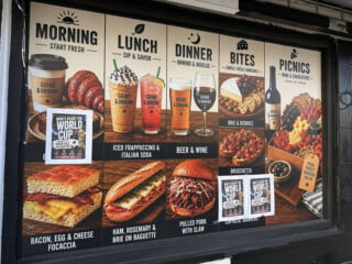 Menu board displaying meal options: breakfast focaccia, Italian soda, baguette sandwich, pulled pork, beer, brie and berries, bruschetta, picnic platters, and World Cup promotion, all with images of the dishes and drinks.