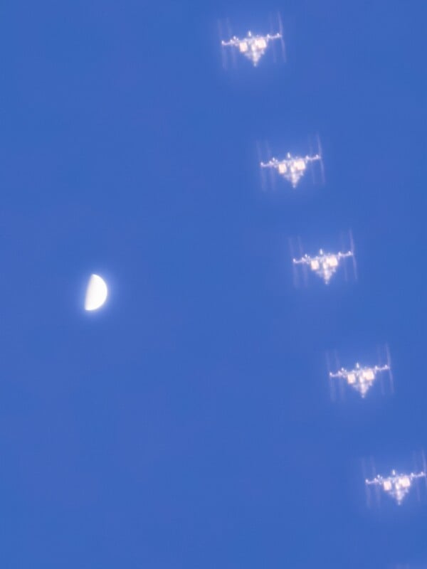 A sequence of the International Space Station passing in front of a half moon in a clear blue sky, captured in multiple exposures to show its movement.