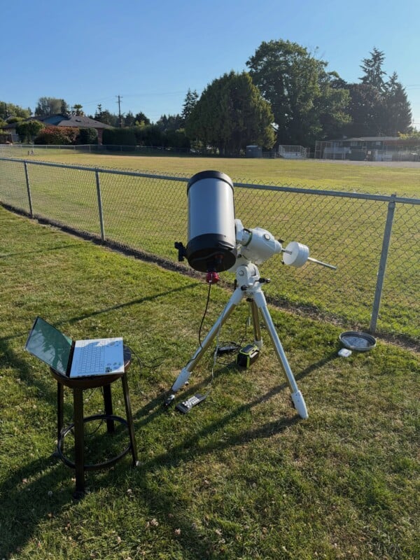 A telescope on a tripod stands on grass near a chain-link fence, with a laptop on a stool beside it. Cables and equipment are on the ground. Trees and houses are visible in the background under a clear sky.