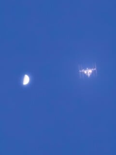 A half moon is visible in a blue sky with the International Space Station appearing nearby, both slightly blurred by atmospheric haze.