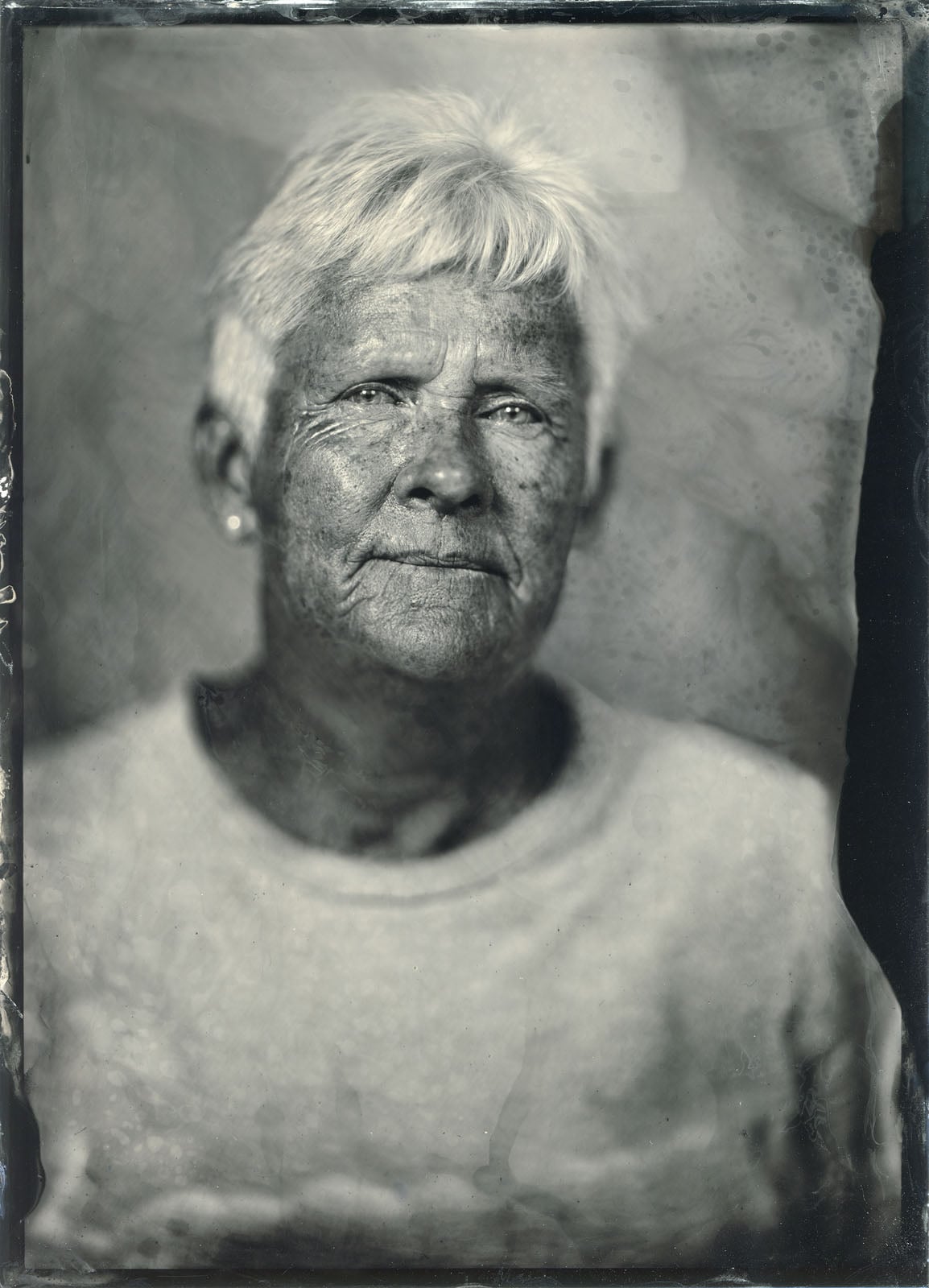 A black-and-white, textured portrait of an older person with short, light hair, wearing a plain shirt against a mottled background.