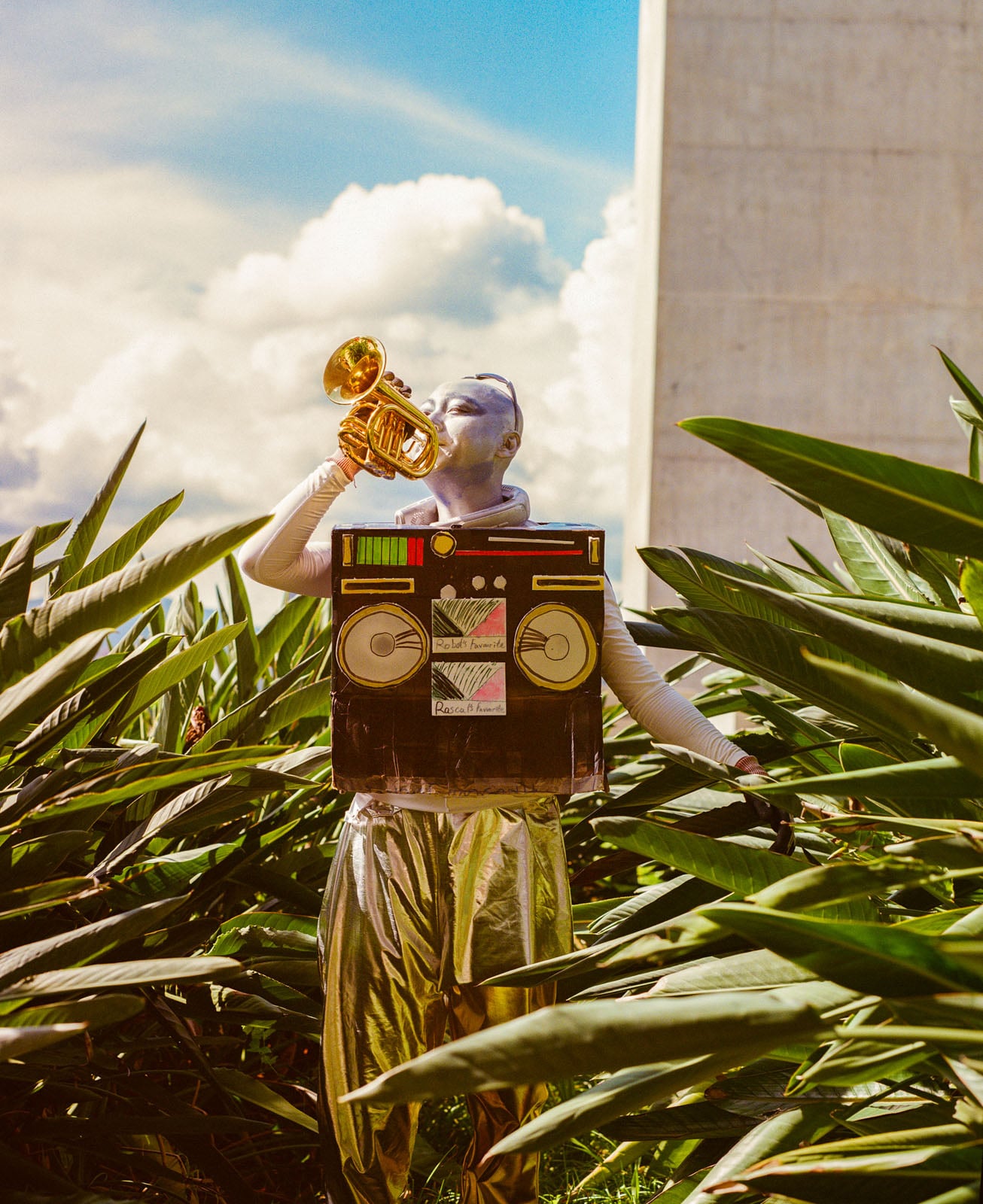 A person painted silver wears a cardboard boombox costume and shiny pants while playing a small brass trumpet in a garden.