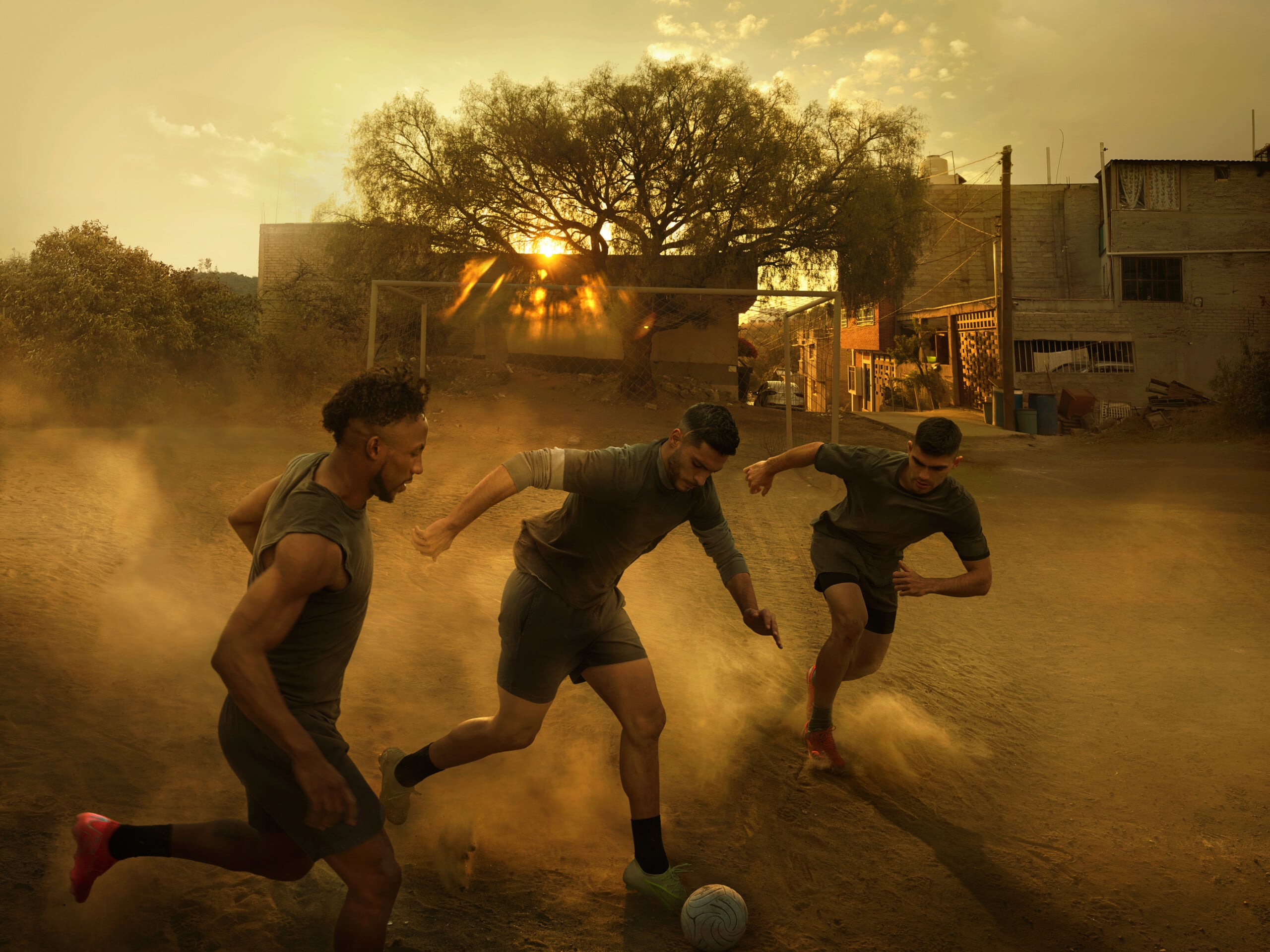 Three men play soccer on a dusty dirt field at sunset, kicking up clouds of dust as they compete for the ball. Houses and a large tree are visible in the background with golden sunlight shining through.