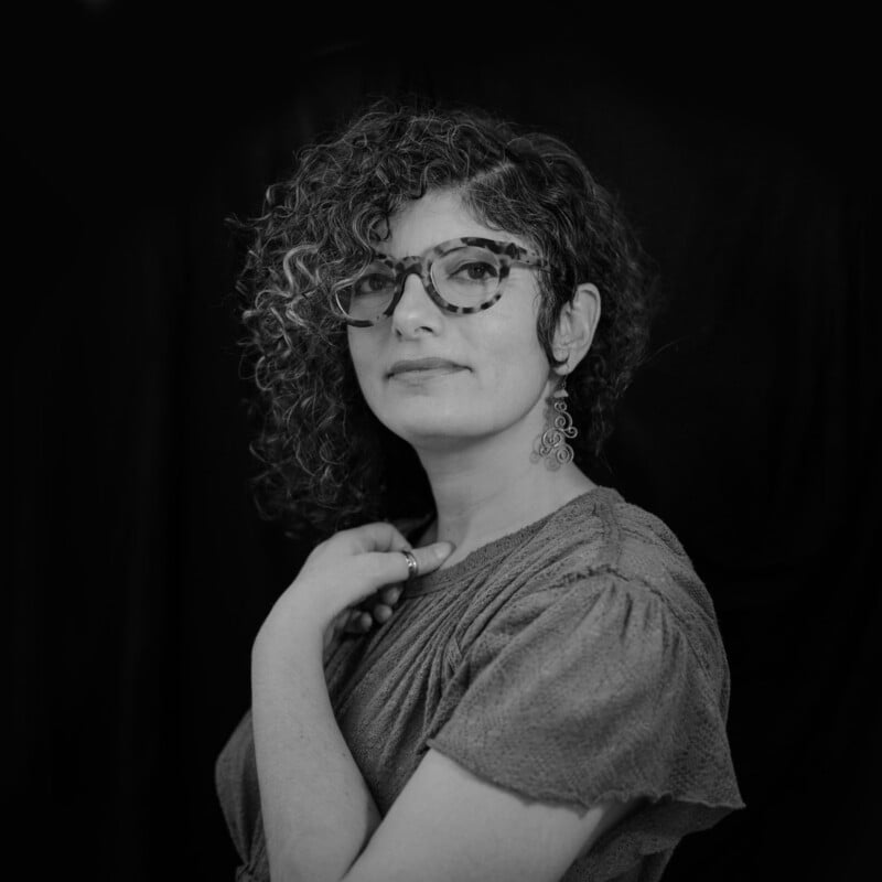 A person with curly hair, glasses, and earrings poses with one hand near their neck, wearing a textured blouse against a dark, plain background.