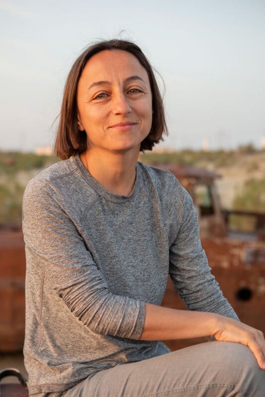 A woman with short brown hair wearing a gray long-sleeve shirt sits outdoors, smiling slightly. The background includes greenery and a rusty, old vehicle under a clear sky.
