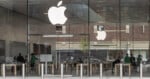 Apple Store with large glass windows, white Apple logos, and several employees in green shirts inside. Wooden tables are arranged in rows, and the outside shows reflections of buildings and trees.