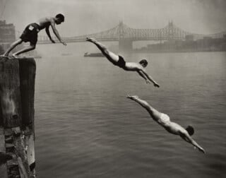Three men dive into a river from a wooden pier, with a large bridge visible in the background.