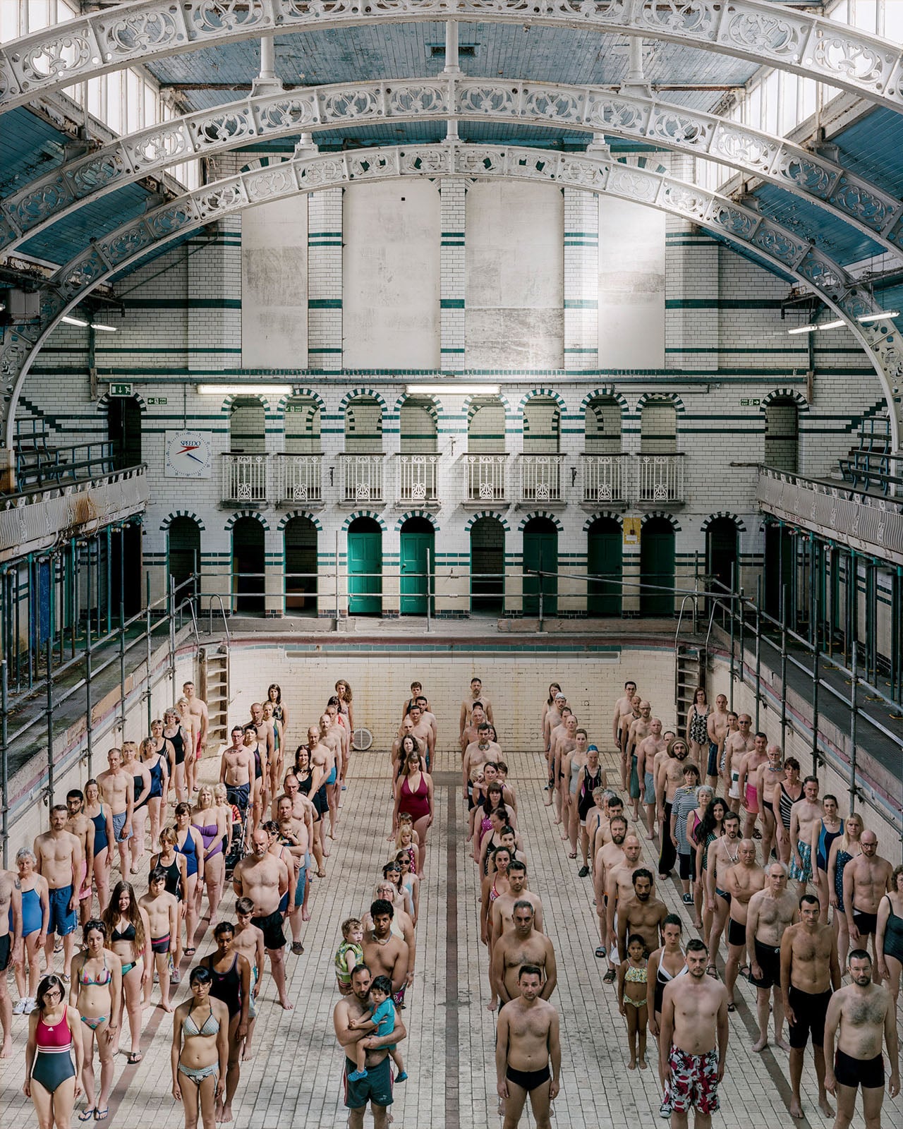 A large group of people stands in rows inside a drained, vintage indoor swimming pool with arched ceilings and tiled walls.