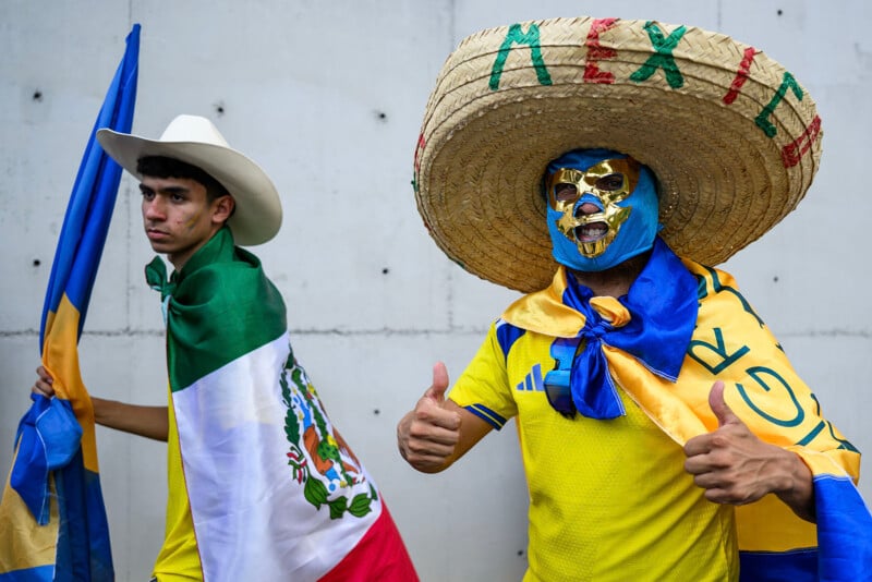 Two men in festive attire pose; one wears a sombrero, lucha libre mask, and gives thumbs up, while the other wears a cowboy hat and is draped in a Mexican flag, both holding colorful flags.