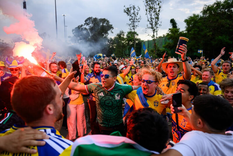 A large crowd of excited soccer fans, mostly in yellow jerseys, rally outdoors. One person holds a flare, while others cheer and wave flags. Trees and a cloudy sky are in the background.