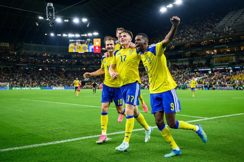 Three soccer players in yellow and blue uniforms celebrate a goal on a stadium field, smiling and raising their arms, with a cheering crowd and scoreboard in the background under bright stadium lights.