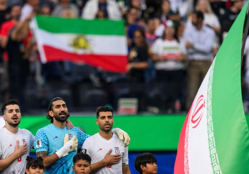 Three Iranian soccer players stand with hands on their chests during a national anthem, with a large Iranian flag in the foreground and a crowd holding another flag in the background.