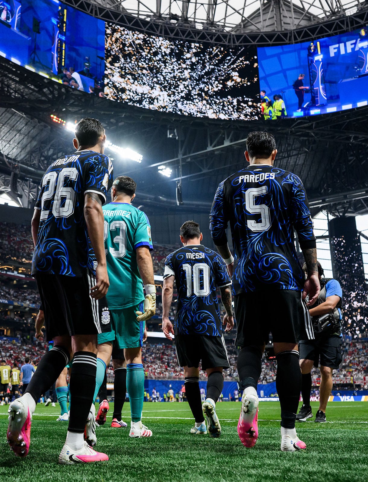 Four soccer players in blue and black jerseys walk onto a stadium field, with confetti flying above and a large crowd in the background. The stadium roof is partially open, letting in light.