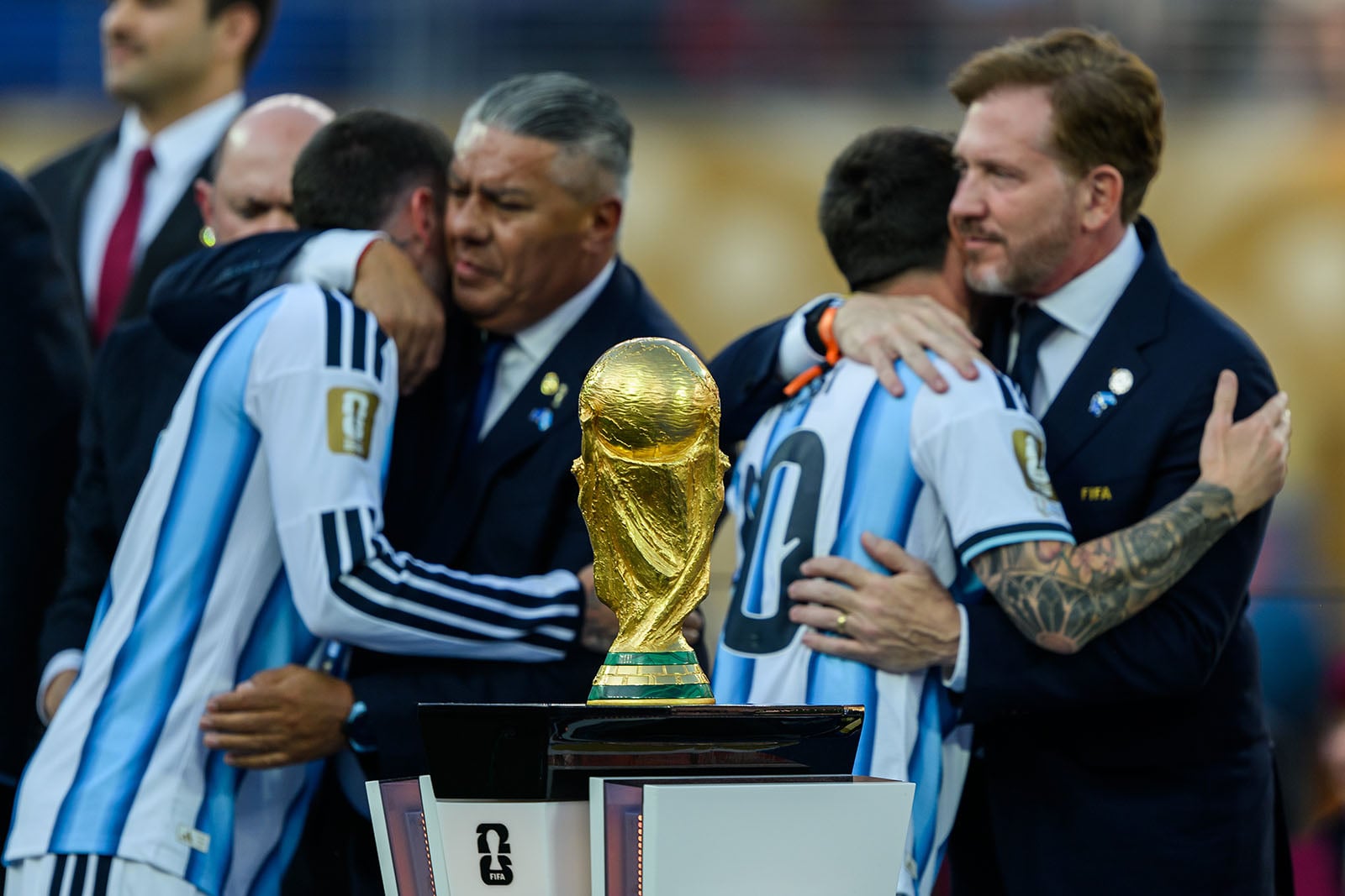 The FIFA World Cup trophy is in focus as members of the Argentina soccer team, wearing blue and white jerseys, embrace and comfort each other in the background.