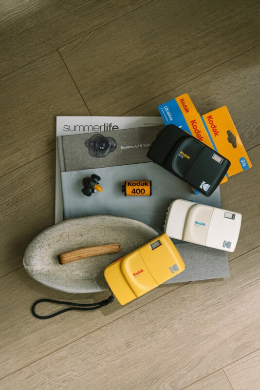 Three Kodak film cameras (yellow, white, black), two Kodak film boxes, a Kodak 400 film roll, earbuds, a lighter, and magazines arranged on a wooden floor.