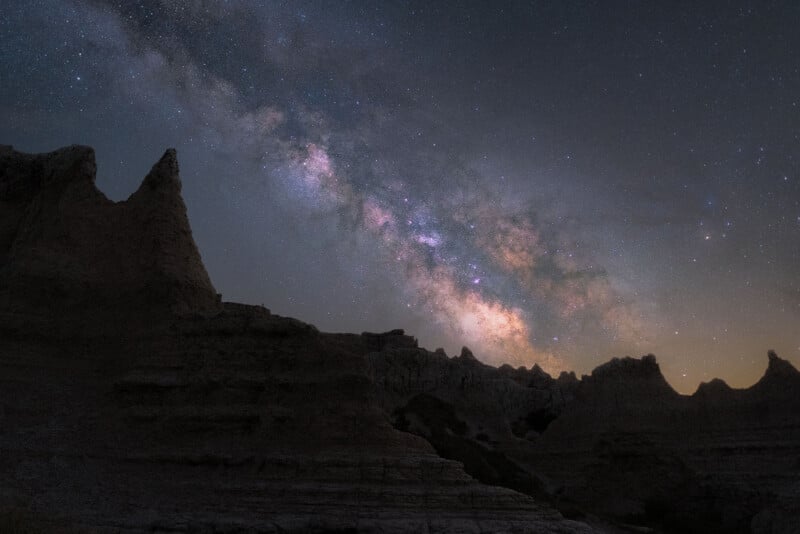 The Milky Way galaxy shines brightly in the night sky above dark, jagged rock formations in a rugged desert landscape.