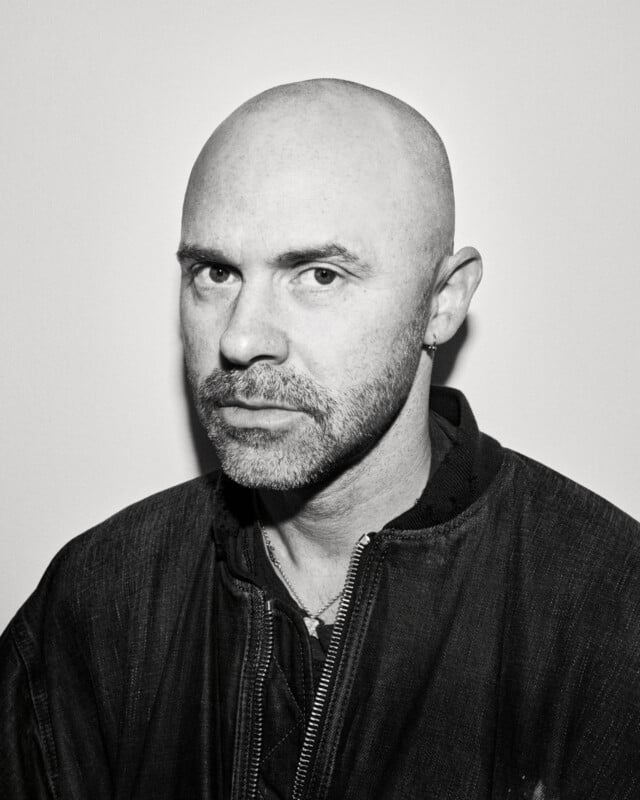 A bald man with a trimmed beard and earring stares seriously at the camera. He wears a dark textured jacket and a necklace, posed against a plain light background in black and white.
