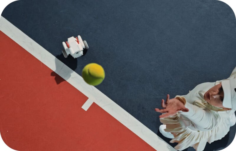 A person in white athletic clothing reaches to catch a tennis ball near a court line, with a small white robot nearby on the court. The image is taken from above, showing red and blue court surfaces.
