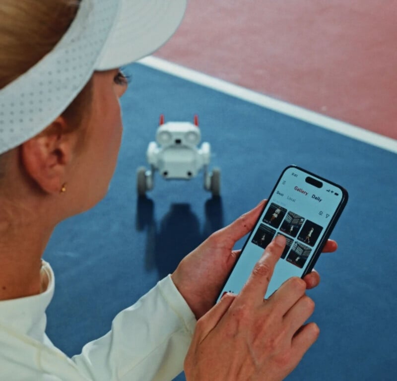 A person wearing a white visor and top uses a smartphone to control a small, white robot dog on a blue and red court. The phone screen displays a gallery of images or videos.
