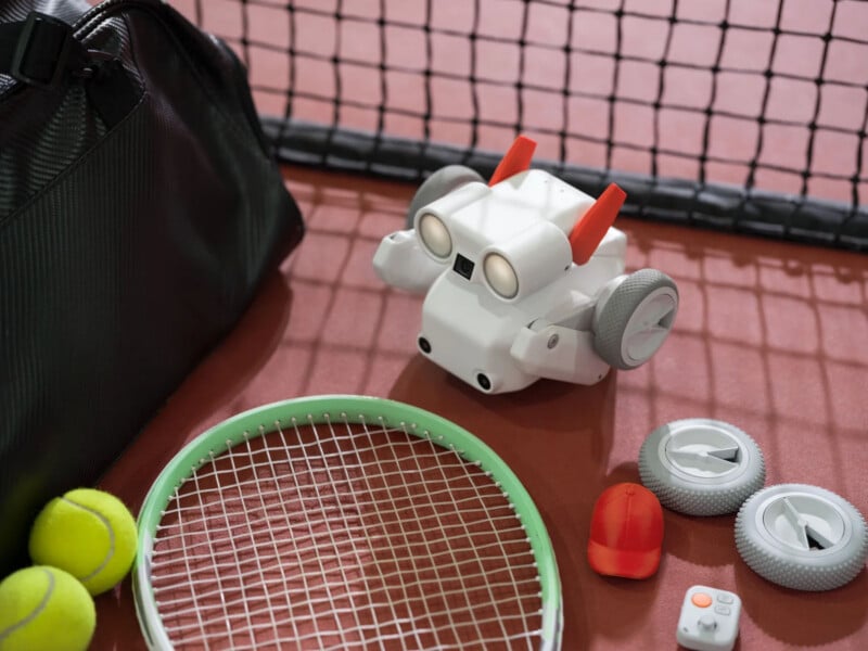 A small white robot with orange accents sits on a tennis court near a black sports bag, tennis racket, tennis balls, extra wheels, a red cover, and a small remote control. A net is visible in the background.