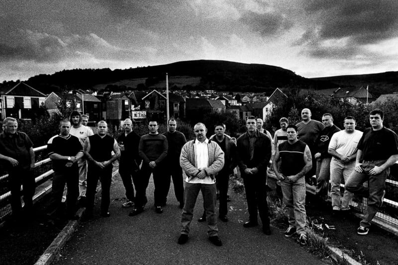 A group of men stand together on a road in front of houses and hills, under a dramatic, cloudy sky; the image is black and white.