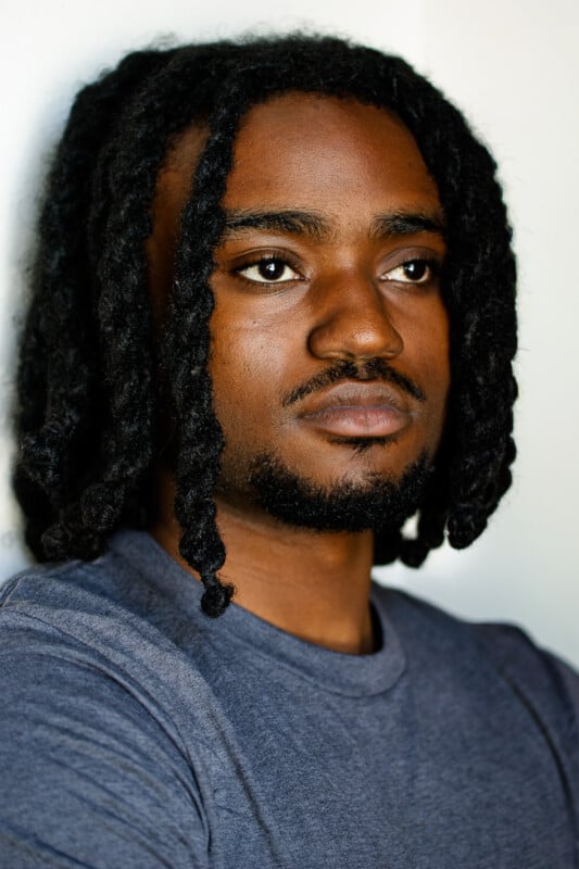 A person with medium-length twisted hair and a short beard is wearing a gray shirt, standing against a light background, looking slightly to the side with a serious expression.