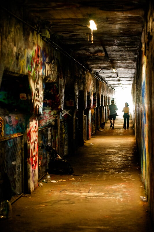 Two people walk toward the light at the end of a long, narrow tunnel covered in colorful graffiti, with dim overhead lights illuminating the passage.