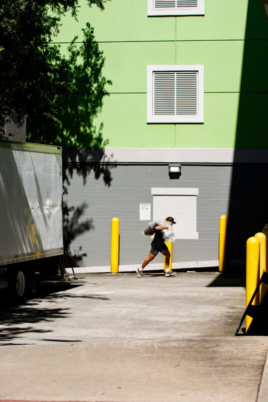 A person in shorts and a cap walks quickly through a sunlit alley, carrying bags near a green and gray building, with yellow posts and a white truck nearby. Shadows partially cover the scene.
