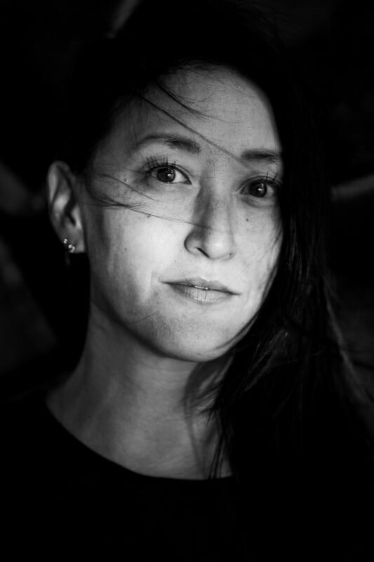 A black and white close-up portrait of a woman with long dark hair, some strands falling across her face. She has a calm expression and is looking directly at the camera. The background is dark and blurred.