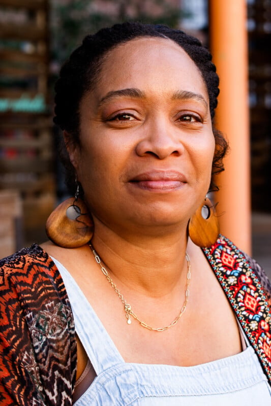 A woman with braided hair, wearing large wooden hoop earrings, a gold chain necklace, and a patterned top, looks at the camera with a calm expression. The background is outdoors and softly blurred.