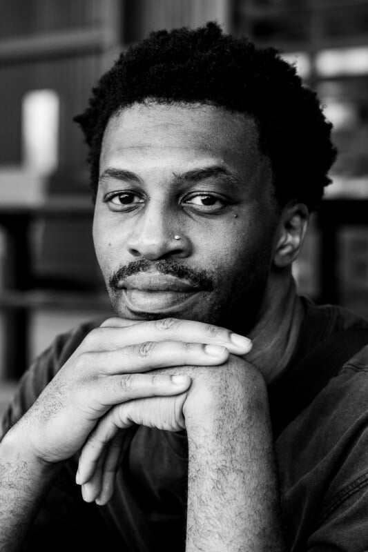 Black and white photo of a man resting his chin on his hands, looking directly at the camera with a calm expression. He has short curly hair and a nose piercing. The background is softly blurred.