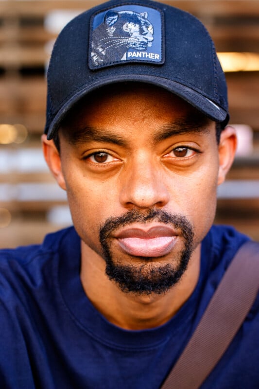 A man with a short beard and mustache wears a black "Panther" cap and a navy blue shirt, looking directly at the camera with a neutral expression. The background is softly blurred.