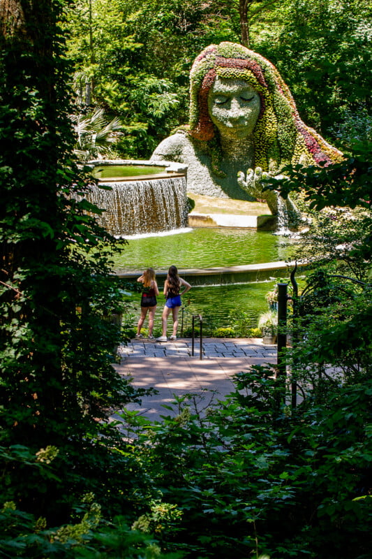 Two people stand in front of a large plant sculpture of a reclining woman with flowing hair beside a waterfall, surrounded by lush greenery in a botanical garden.
