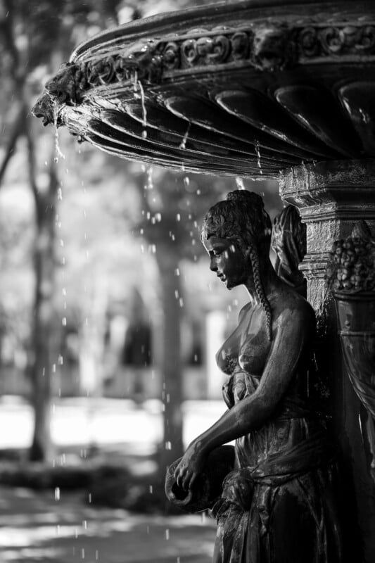 Black and white photo of a decorative fountain featuring a detailed statue of a woman with braids, water dripping from the fountain above her, and a blurred outdoor background.